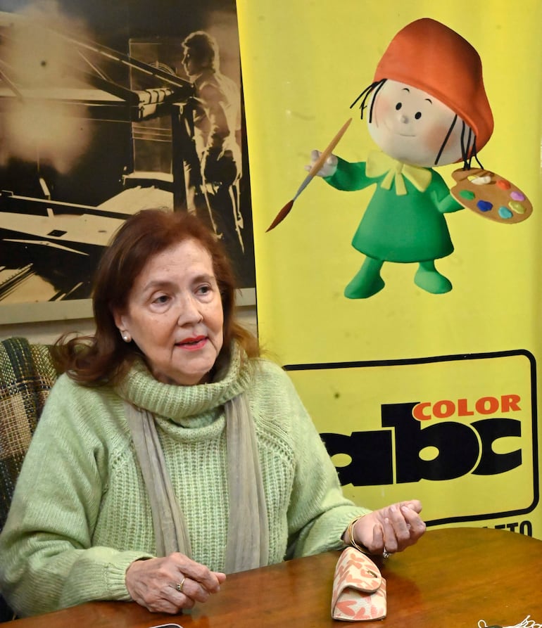 Stael Ruffinelli de Ortiz sentada con suéter verde claro y chal gris, gesticulando hacia un objeto en la mesa.