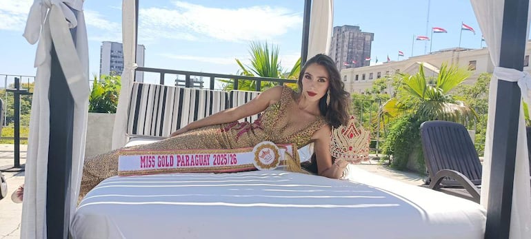 Mujer sentada en cama con vestido dorado, corona y banda de Miss Gold Paraguay, rodeada de banderas de Paraguay en el fondo.