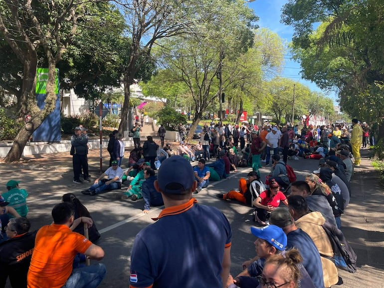 Decenas de personas sentadas, algunas vestidas con camisetas de colores, escuchando un orador en un parque urbano.
