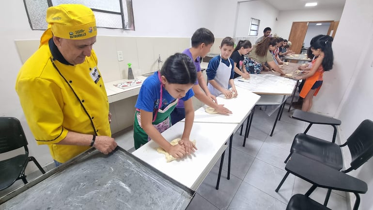 Instructor en delantal amarillo observa a niños con delantales variados amasando masa en mesas de cocina.