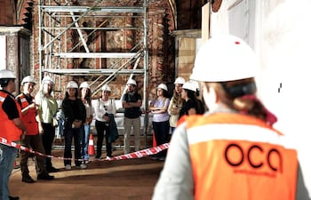 Varios trabajadores con cascos prestan atención a un guía en la restauración de un histórico templo, rodeados de andamios.