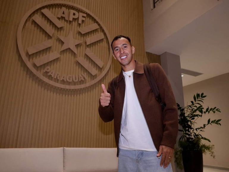 Alejandro Romero Gamarra Kaku sonríe con pulgar hacia arriba, vistiendo abrigo marrón y camiseta blanca, frente al logo de la APF.