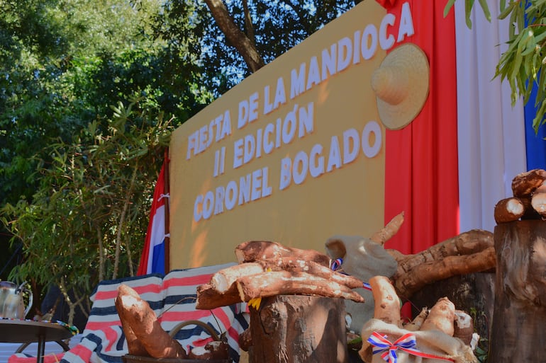 Escenario decorado con raíces de mandioca y sombrero de paja, rodeado de vegetación, para la Fiesta de la Mandioca.