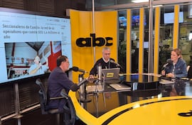Tres personas en estudio de televisión: hombre con gafas y camisa azul habla al micrófono, otro hombre con suéter negro escucha, mujer con chaqueta de mezclilla sostiene un vaso.