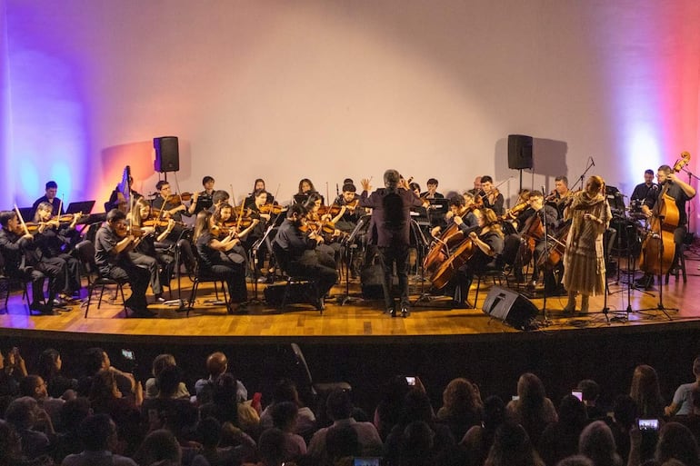 Director de orquesta en vestimenta oscura dirige a músicos con instrumentos de cuerda, mientras el público graba el evento con móviles.