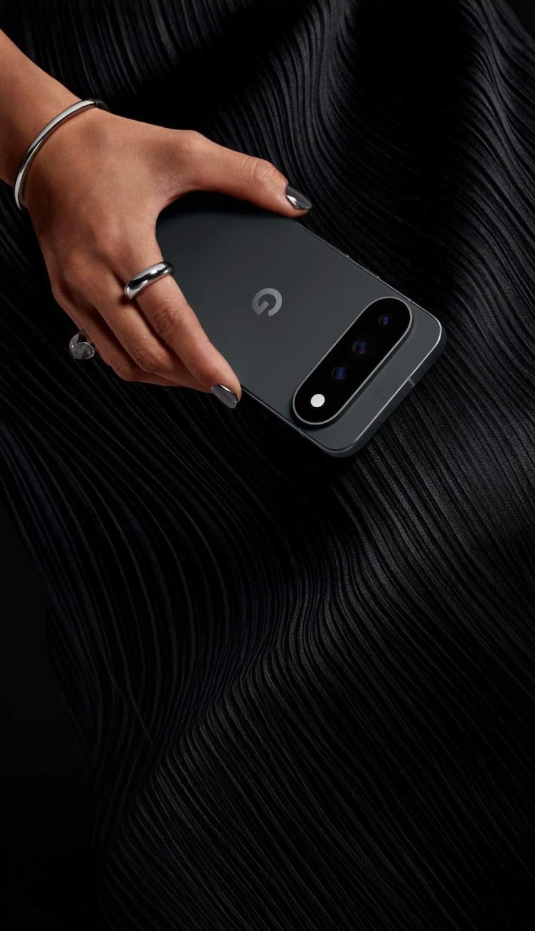 Mano con esmalte negro y anillos sostiene un teléfono gris oscuro, sobre fondo negro texturizado.