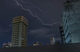 Paisaje urbano nocturno con rayo iluminando el cielo y edificios reflejando luces en un ambiente oscuro.