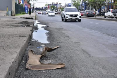 Calle inundada con vehículos, pavimento dañado y bache visible, bajo un cielo nublado en un entorno urbano.