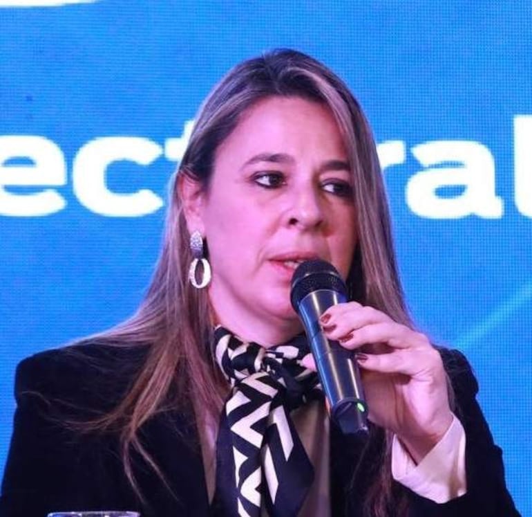Mujer de cabello largo y suelto, con conjunto oscuro y bufanda de rayas, hablando al micrófono frente a fondo azul.