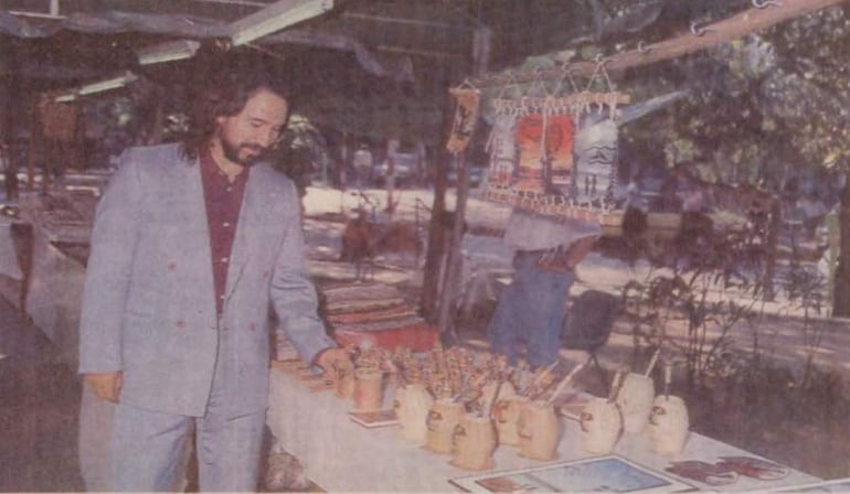 Marco Antonio Solís, vestido con traje claro, contempla máscaras y artesanías en un vibrante mercado al aire libre.