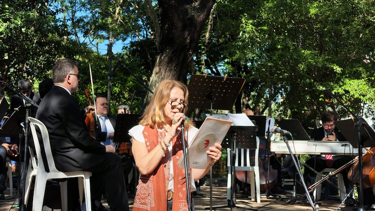 María de la Paz Rivarola con micrófono, rodeado de músicos en traje oscuro en un parque con árboles.