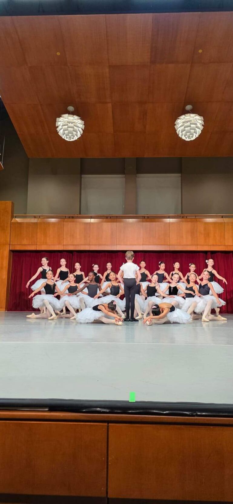 Bailarinas en tutús blancos y camisetas negras, algunas de pie y otras en el suelo, bajo luces de un escenario con cortinas rojas.