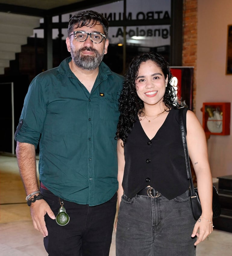 Hombre de cabello corto y barba canosa en camisa verde, junto a mujer de cabello rizado en blusa negra, posan sonriendo en un ambiente moderno.