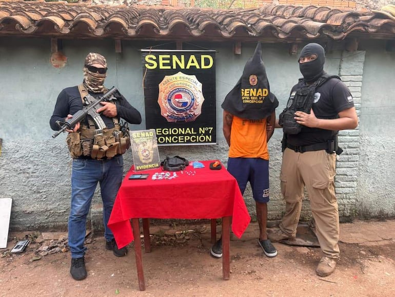 Agentes de la SENAD con vestimenta táctica y un detenido con capucha, en un ambiente gris y con una mesa roja frente a ellos.