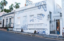 Dos personas conversan cerca de un mural blanco y azul, mientras una tercera pinta en el suelo. Ambiente creativo y colaborativo.