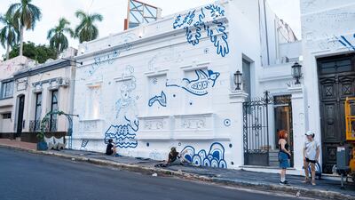 Dos personas conversan cerca de un mural blanco y azul, mientras una tercera pinta en el suelo. Ambiente creativo y colaborativo.
