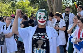 Persona con máscara de 'Joker' y bata blanca levanta el puño junto a otros médicos, en un ambiente de protesta con una bandera.
