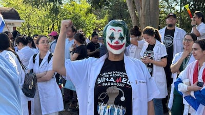 Persona con máscara de 'Joker' y bata blanca levanta el puño junto a otros médicos, en un ambiente de protesta con una bandera.