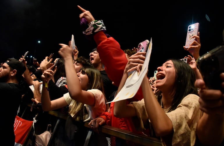 Multitud en concierto de Milo J, con manos levantadas, sosteniendo teléfonos y carteles. Atmosfera vibrante y luces de colores.