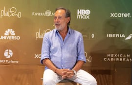 Guillermo Francella, de camisa azul clara y pantalones blancos, escucha atentamente en una sala decorada con logotipos de marcas.