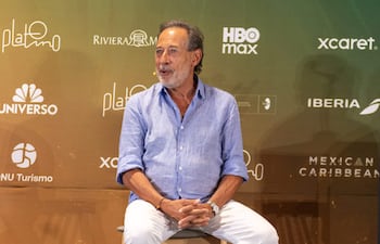 Guillermo Francella, de camisa azul clara y pantalones blancos, escucha atentamente en una sala decorada con logotipos de marcas.