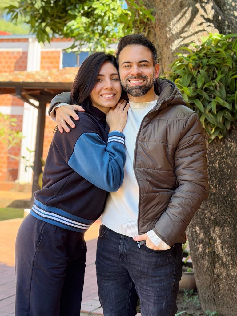 Silvia Romero con conjunto deportivo oscuro y azul abrazando a David Mussi, que lleva abrigo marrón, ambos sonrientes en un jardín.