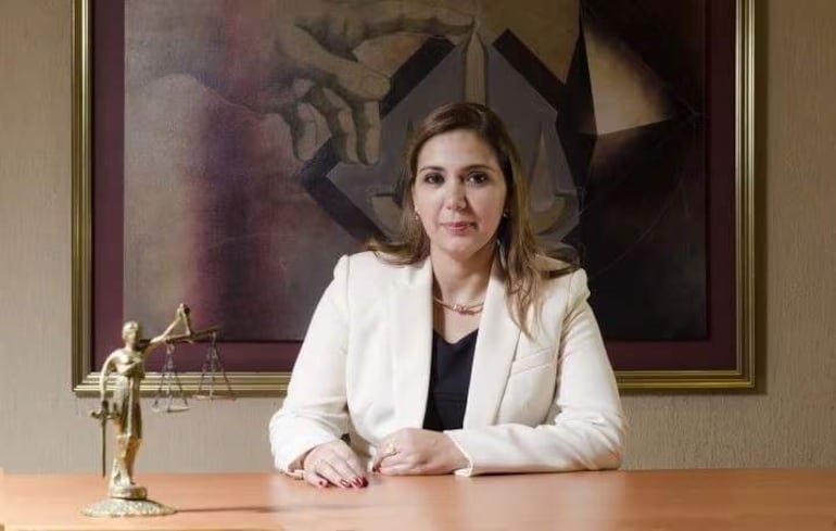 Nancy Graciela Salomón Marín con blusa negra y blazer blanco, en su oficina con figura de la Justicia y pintura abstracta.