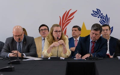 Mujer de cabello rubio con lentes habla al micrófono, mientras cinco hombres la observan serios, sentados en una mesa cubierta con mantel negro.