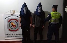 Dos hombres encapuchados y esposados en un entorno policial con un oficial de policía observando.