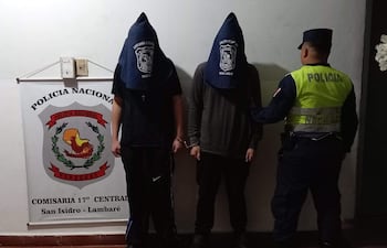 Dos hombres encapuchados y esposados en un entorno policial con un oficial de policía observando.
