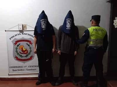 Dos hombres encapuchados y esposados en un entorno policial con un oficial de policía observando.