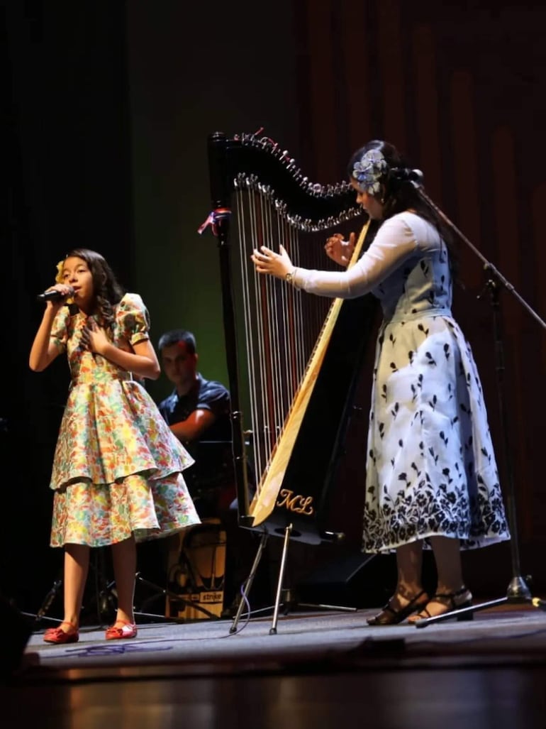 Niña en vestido colorido canta con micrófono, acompañada de mujer con arpa en escenario iluminado.