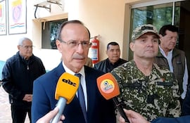 Óscar González con traje oscuro y corbata, sostiene un micrófono, mientras un militar en uniforme lo acompaña en un edificio público.