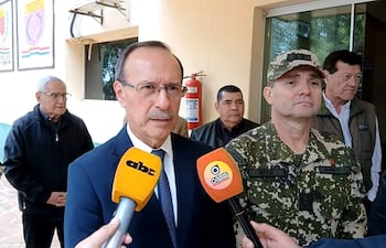 Óscar González con traje oscuro y corbata, sostiene un micrófono, mientras un militar en uniforme lo acompaña en un edificio público.