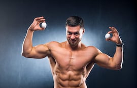Hombre musculoso con torso desnudo sostiene un huevo en cada mano, mostrando confianza en un fondo oscuro.