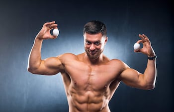 Hombre musculoso con torso desnudo sostiene un huevo en cada mano, mostrando confianza en un fondo oscuro.