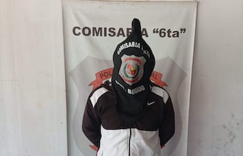 Hombre con abrigo blanco y negro y capucha negra, de pie en una comisaría, mirando al frente en un ambiente austero.