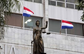 Escultura femenina de la Justicia sosteniendo balanzas, con banderas de Paraguay ondeando en el fondo.