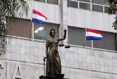 Escultura femenina de la Justicia sosteniendo balanzas, con banderas de Paraguay ondeando en el fondo.