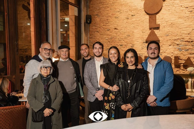 Grupo de ocho personas sonrientes en un café con paredes de ladrillo, destacando una mujer con abrigo oscuro y un hombre canoso con gafas.