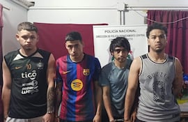 Cuatro hombres de pie, con camisetas diversas, alineados y esposados, en un entorno de fondo burdeos con insignias policiales.