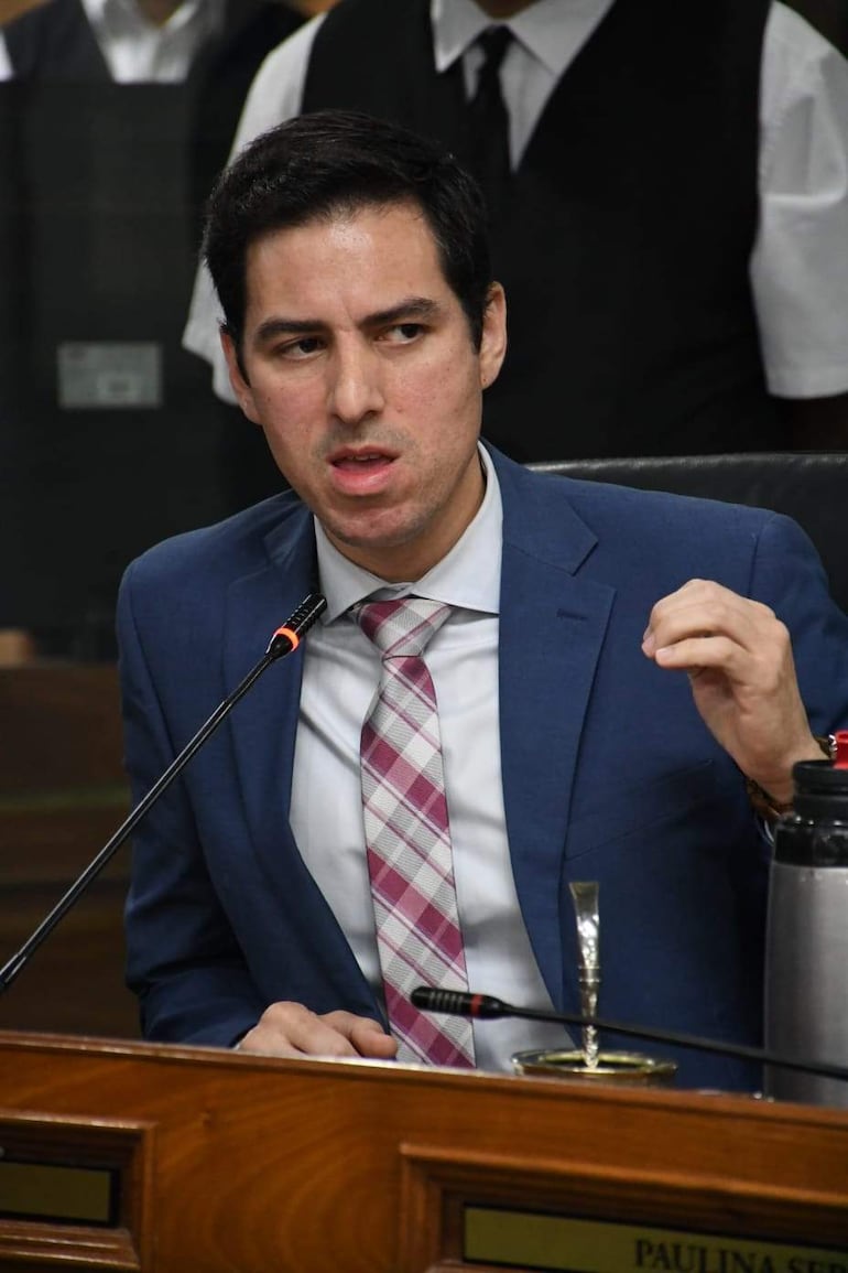 Álvaro Grau sentado al frente con traje azul y corbata de rayas, hablando enfáticamente en un ambiente formal con asistentes atentos.