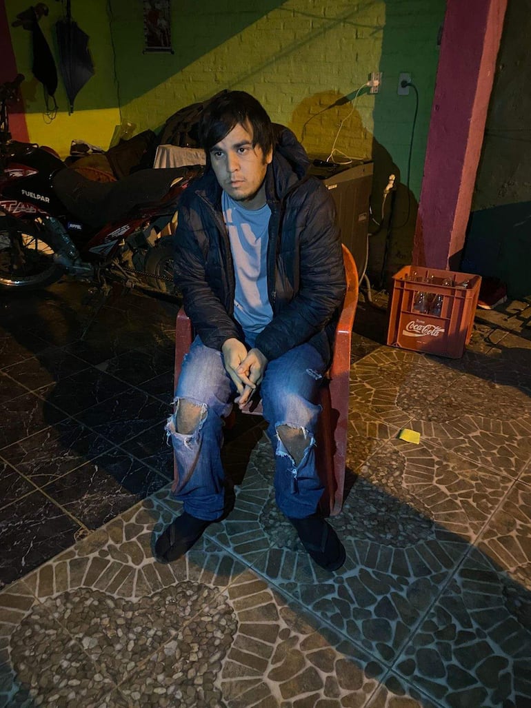 Hombre sentado en silla roja, chaqueta azul oscura y camiseta clara, pensativo en ambiente exterior con luz suave.