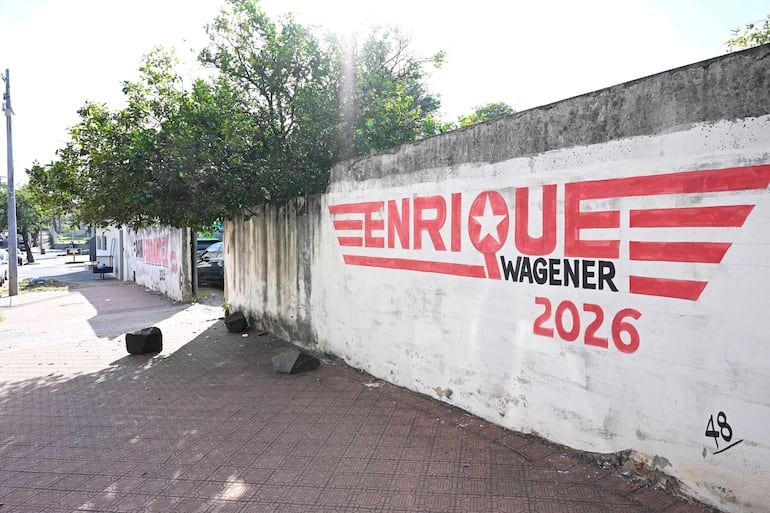 Mural en pared con inscripción 'ENRIQUE WAGENER 2026', rodeado de acera y árboles, sin personas presentes.