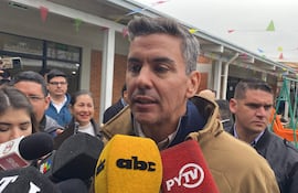 Santiago Peña con abrigo ligero, sosteniendo un micrófono amarillo, rodeado de reporteros en un ambiente festivo.