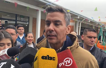Santiago Peña con abrigo ligero, sosteniendo un micrófono amarillo, rodeado de reporteros en un ambiente festivo.