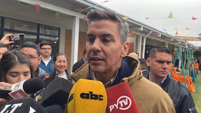 Santiago Peña con abrigo ligero, sosteniendo un micrófono amarillo, rodeado de reporteros en un ambiente festivo.