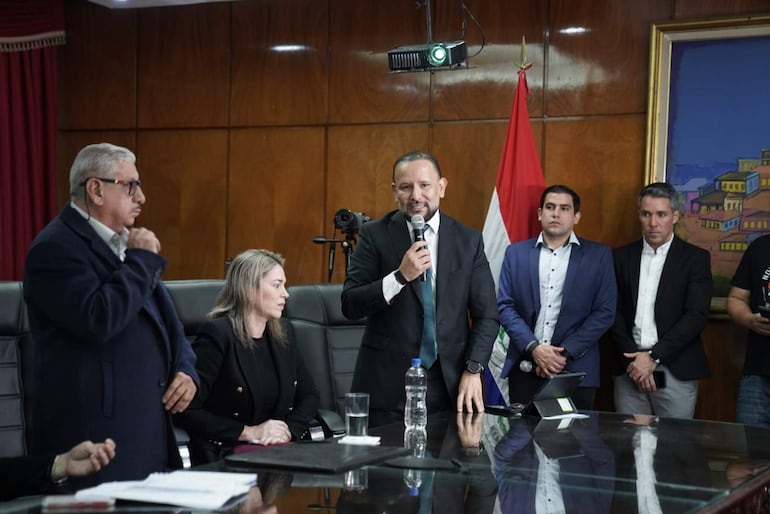Isaías Fretes con micrófono, vestido formal, mientras Cecilia Rodríguez observa atentamente, rodeado de otros hombres en un ambiente formal.