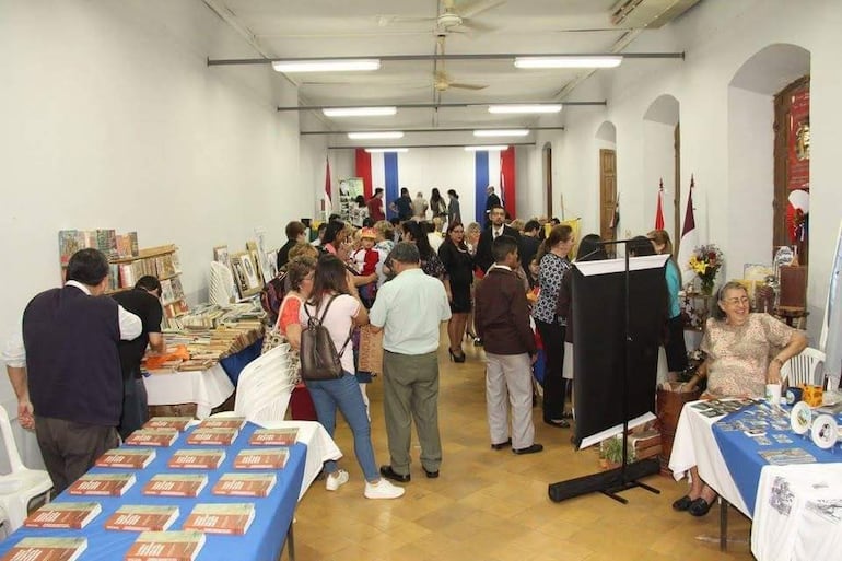 Personas conversando y examinando exhibiciones de libros y productos en un salón iluminado, con banderas decorativas al fondo.