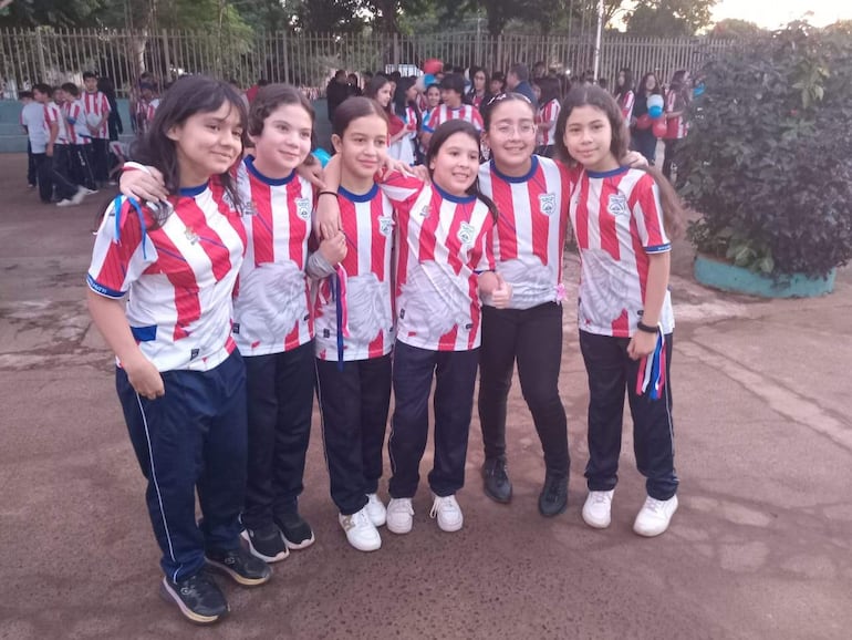 Seis niñas sonrientes en camisetas rojas y blancas, posando alegremente en un evento al aire libre, con vegetación cercana.